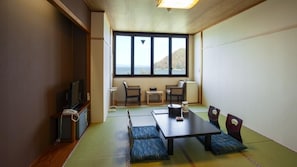 A room with tatami flooring, a low table with cushions, a television, and a window with a view of the sea.