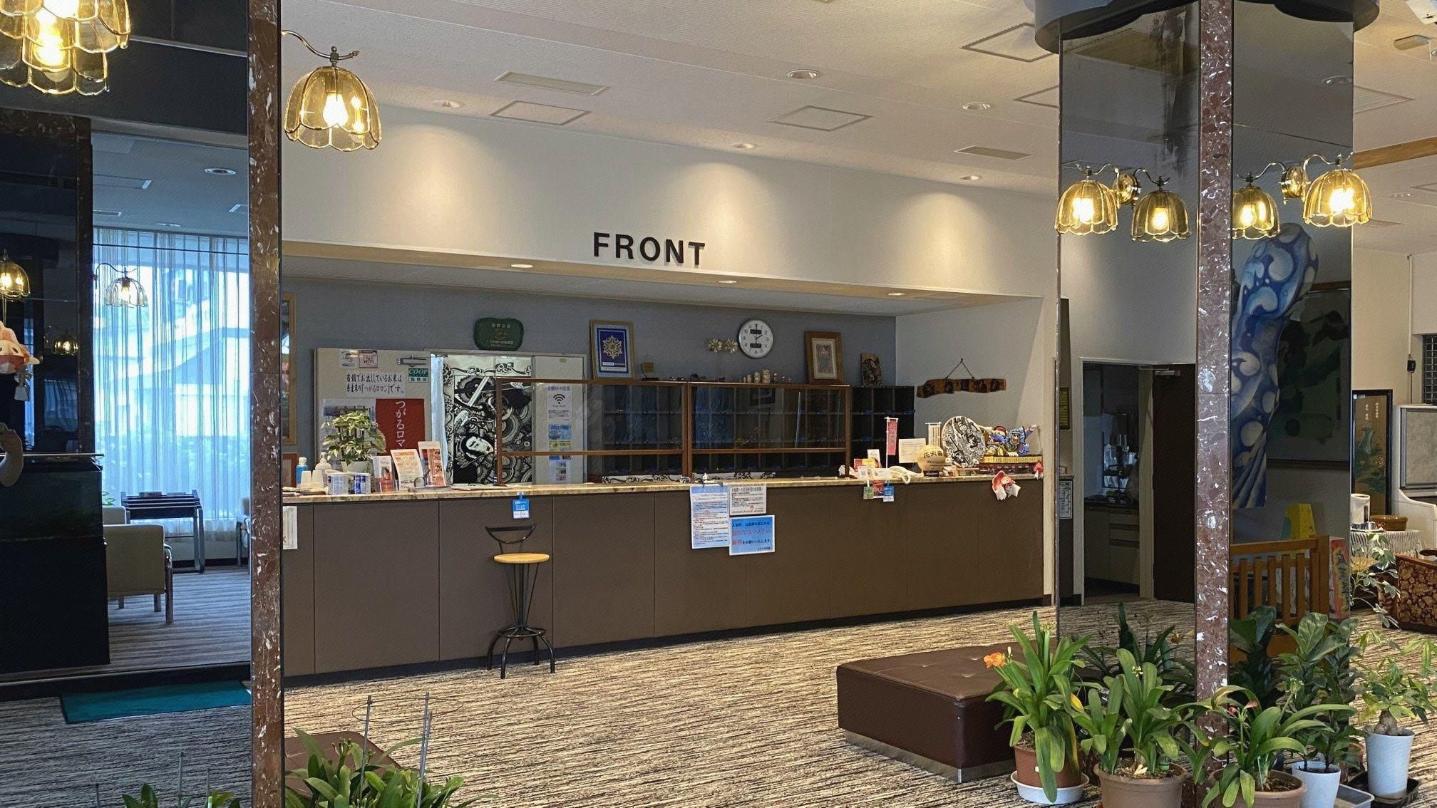 A reception desk labeled 'FRONT' with a clock, informational posters, and a counter with various items. There are potted plants and a seating area with a chair.
