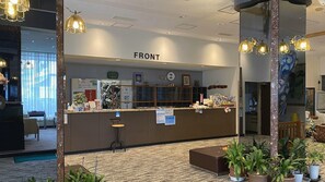 A reception desk labeled 'FRONT' with a clock, informational posters, and a counter with various items. There are potted plants and a seating area with a chair. - Ryokan Asamushi Onsen Hotel Akitaya (Aomori)