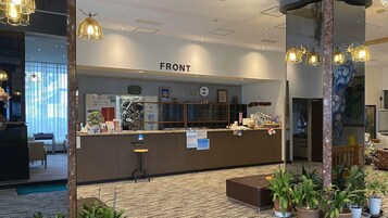 Bancada de recepção com a placa 'FRONT', um relógio, cartazes informativos e um balcão com diversos objetos. Há plantas em vasos e uma área de estar com uma cadeira.