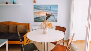 A dining area with a round table, chairs, a wooden bench, a wall-mounted lamp, and a painting of a boat on a pier. - Marina 3 - One Bedroom Apartment, Sleeps 4 (Roses)