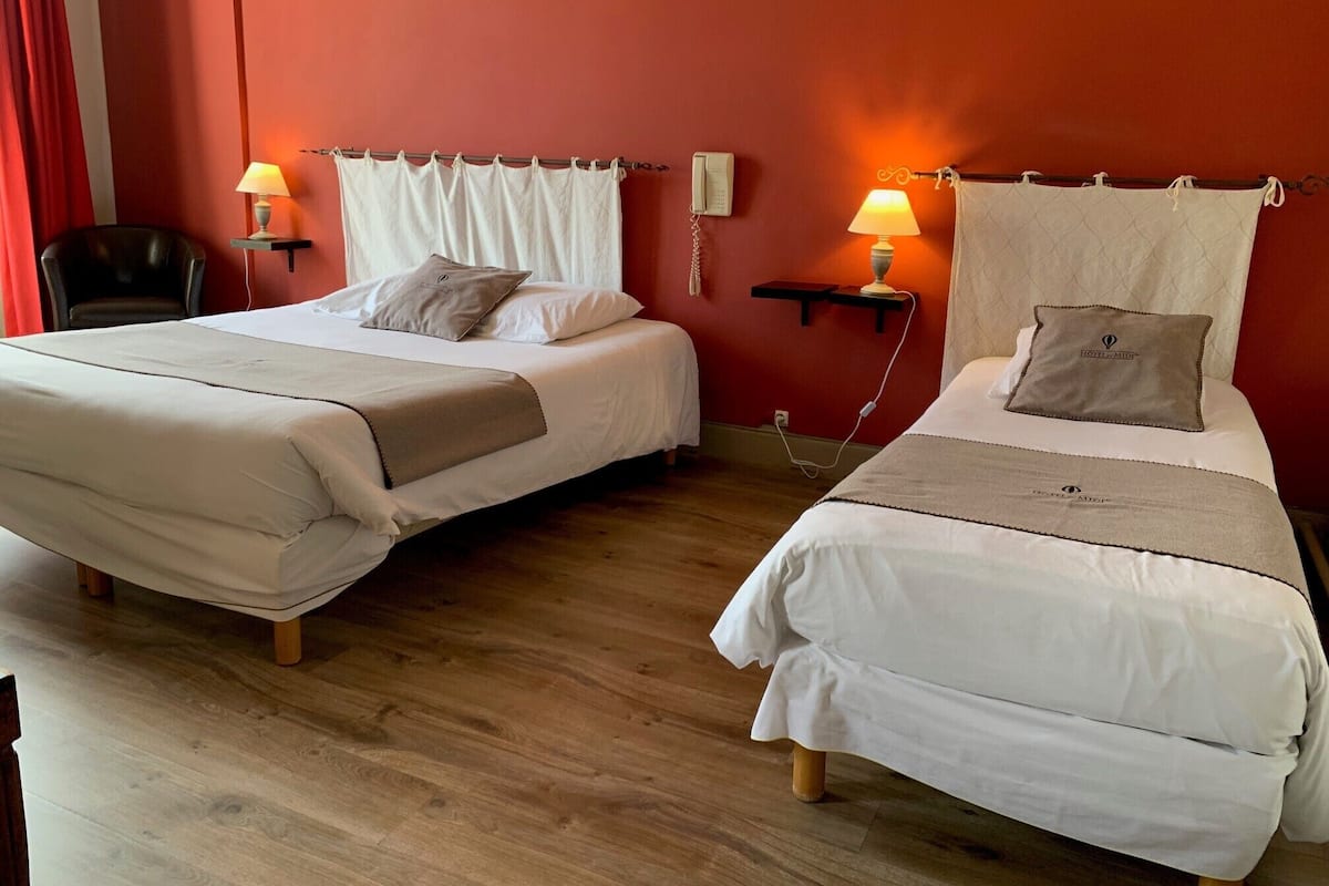 A hotel room with two beds, each with a pillow and a bedside lamp. There is a chair and a small table with a lamp.
