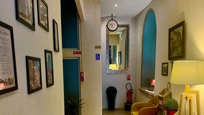 A hallway with framed pictures on the wall, a clock, and a mirror. There are chairs and a table with a lamp. - Hotel du Midi (Annonay)