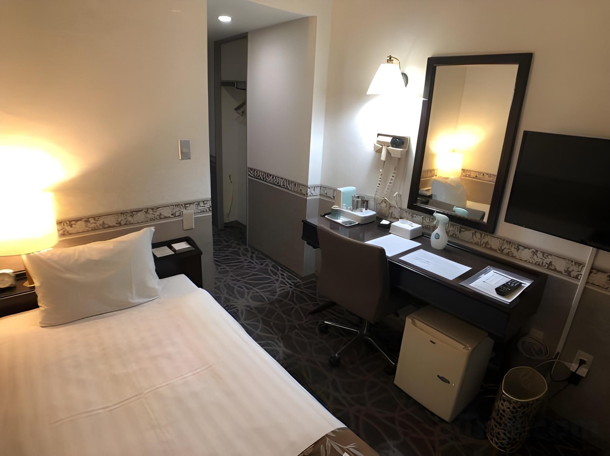 Habitación de hotel con cama, escritorio, silla, refrigerador pequeño, lavamanos, espejo y lámpara.