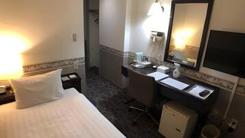 Habitación de hotel con cama, escritorio, silla, refrigerador pequeño, lavamanos, espejo y lámpara.