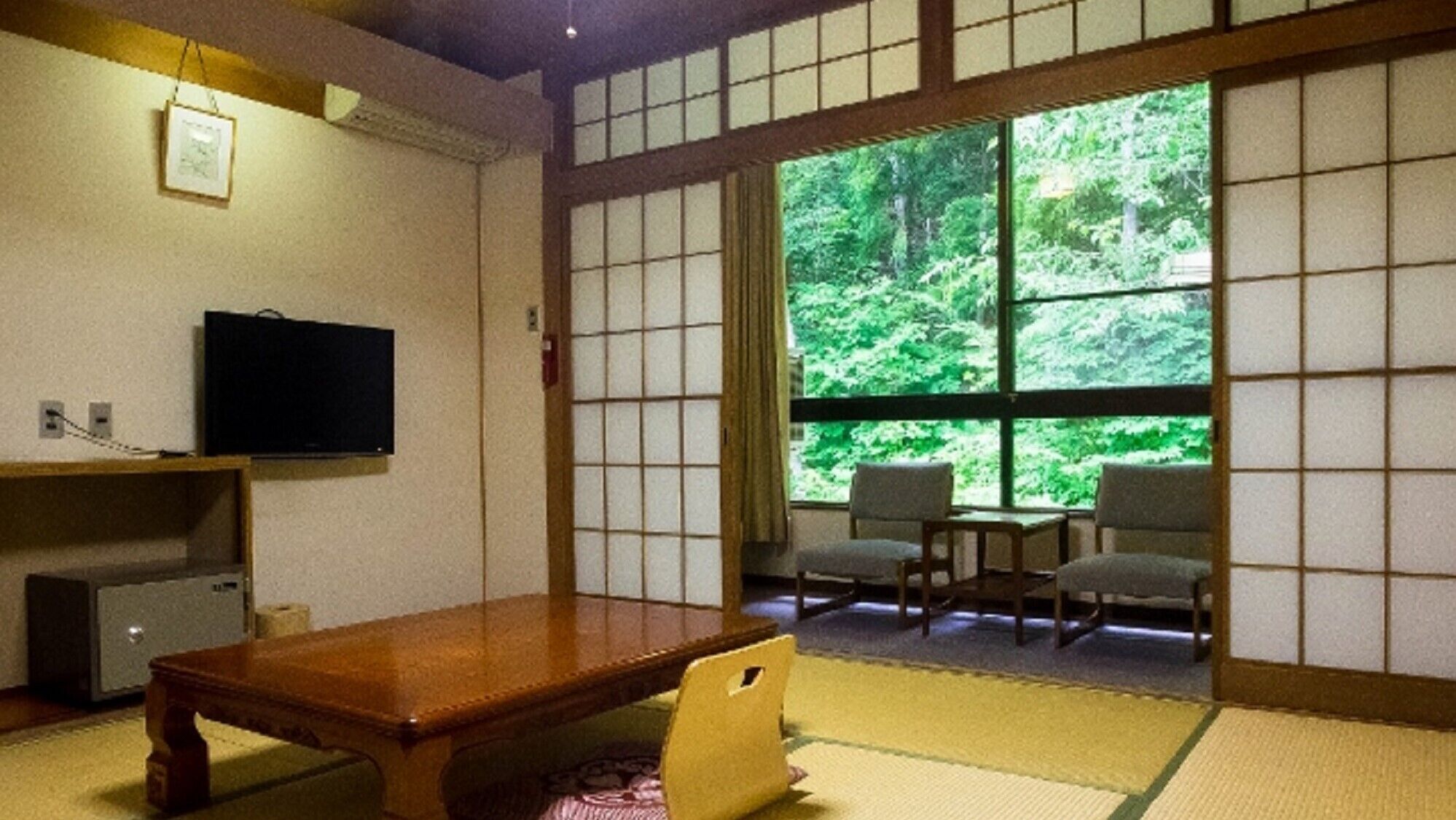 Una habitación japonesa tradicional con suelo de tatami, una mesa de madera baja, un televisor y vistas a un bosque a través de ventanas amplias.