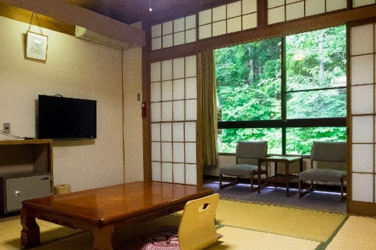 Une pièce de style japonais traditionnel, avec un sol recouvert de tatamis, une basse table en bois, un téléviseur et une vue sur une forêt, visible à travers de grandes fenêtres.