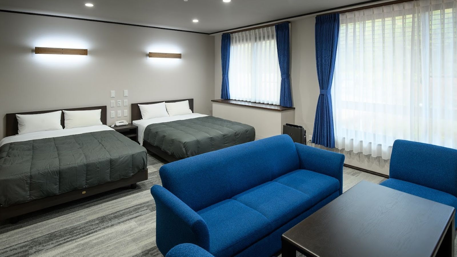 Habitación de hotel con dos camas, un sofá azul, una mesa y un ventanal con cortinas.