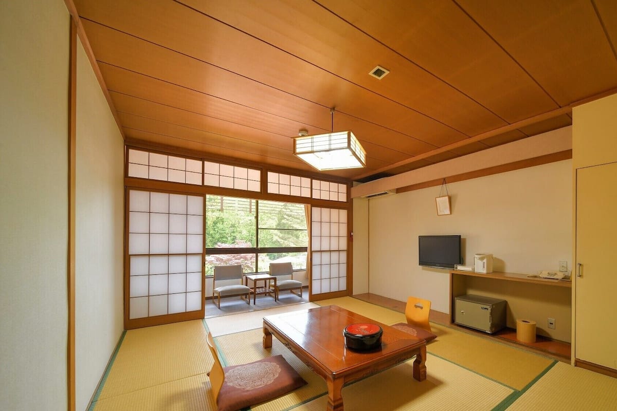 Une pièce de style japonais traditionnel, avec un sol recouvert de tatamis, une petite table sur laquelle se trouve un objet rouge, un téléviseur et un meuble.