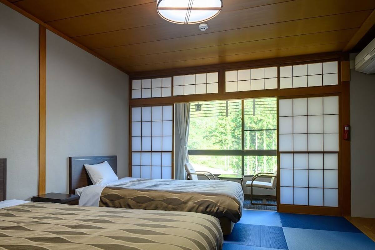 Une chambre d’hôtel avec deux lits, une vue sur la verdure grâce à de grandes fenêtres et une porte coulissante de style japonais traditionnel.