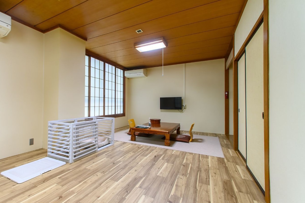 A room with a wooden floor, a white crib, a wooden table with chairs, a television mounted on the wall, and a sliding door.