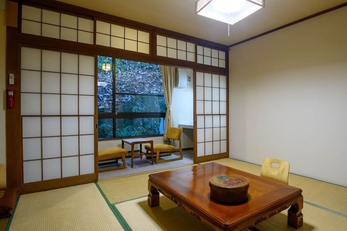 Une pièce de style japonais traditionnel, avec un sol recouvert de tatamis, une petite table ornée d’un objet décoratif et des portes coulissantes donnant sur un paysage verdoyant.