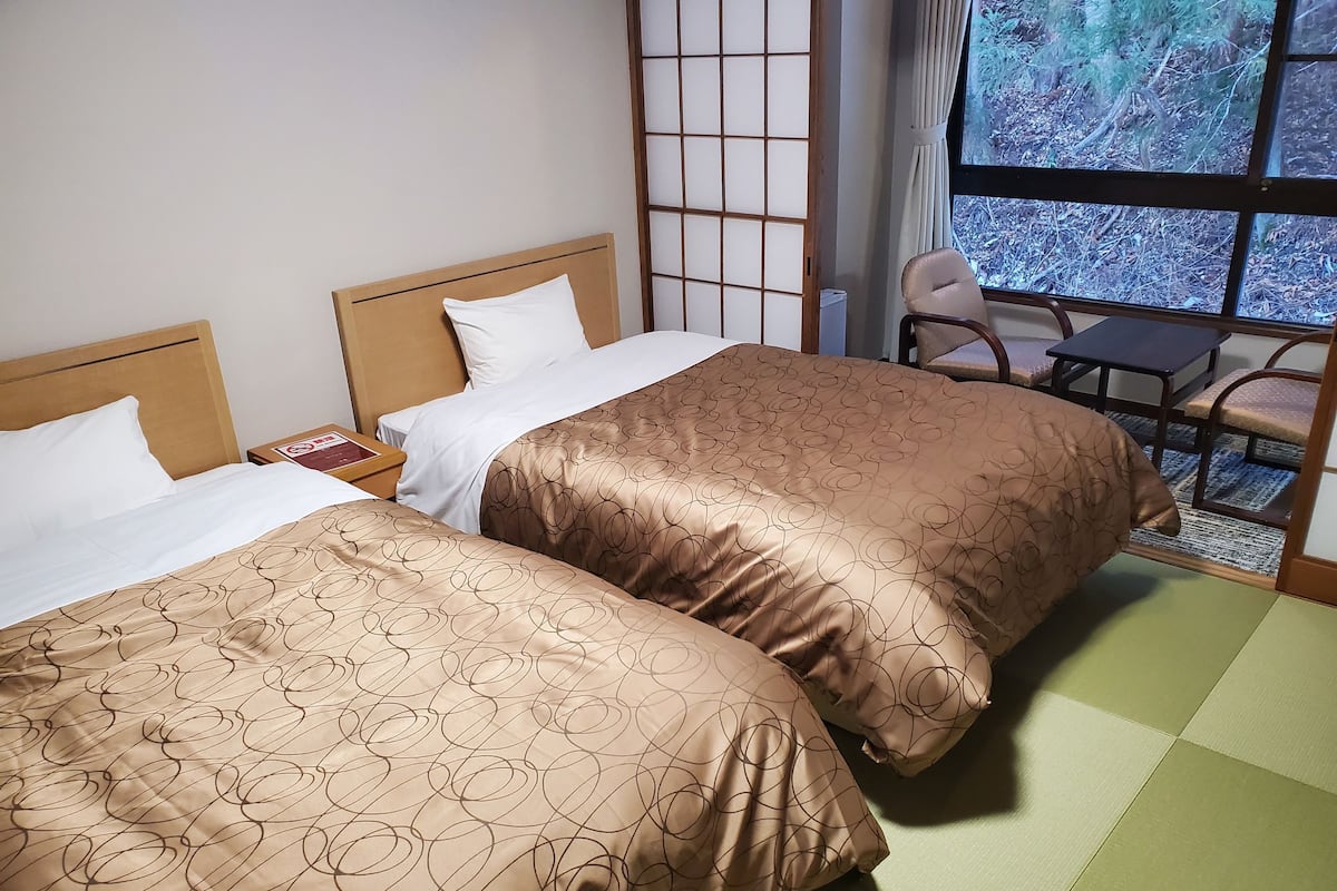 Une chambre d’hôtel avec deux lits, une chaise, une table de chevet et une fenêtre donnant sur des arbres.