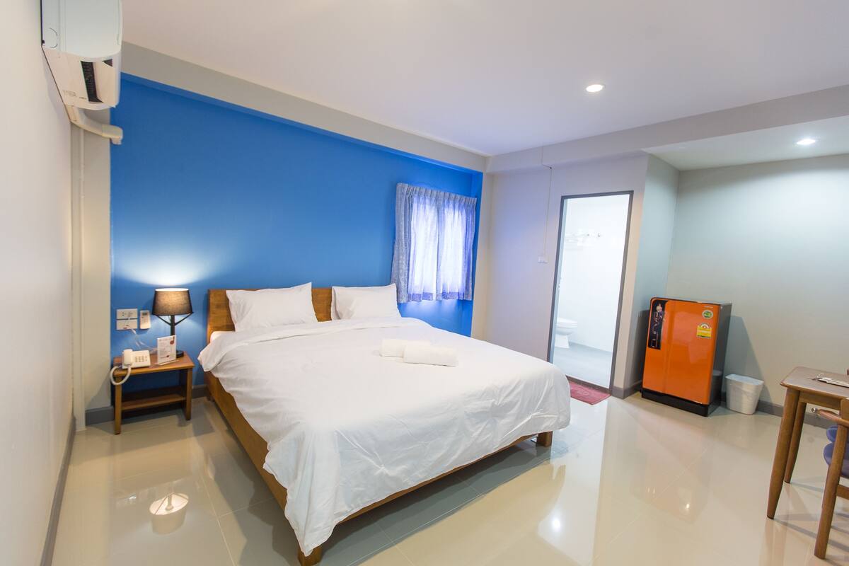 A hotel room with a large bed, a nightstand with a lamp, a refrigerator, and a wooden table.