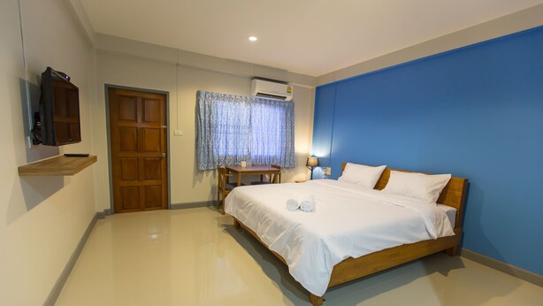 A bedroom with a bed, a wooden nightstand, a table with a lamp, a TV mounted on the wall, and an air conditioning unit. - S3 Room (Sattahip)