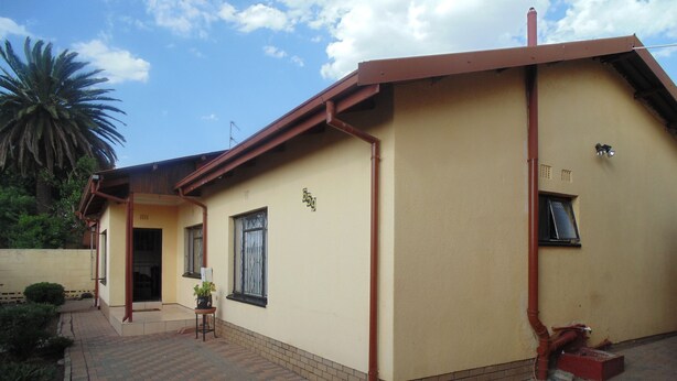 A two-story building with a yellow exterior, a brick pathway leading to the entrance, and a small garden area with a palm tree. - TDJ's B&B (Soweto)