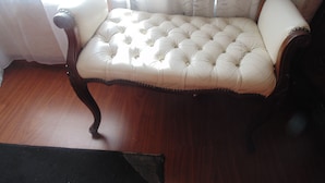 A tufted sofa with wooden armrests and legs, positioned in a room with wooden flooring and a window with sheer curtains. - TDJ's B&B (Soweto)
