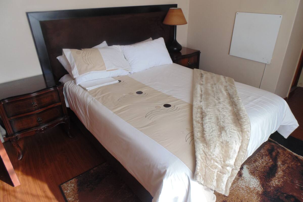 Una cama bien tendida con cabecera, una lámpara de noche y una pizarra en la pared.