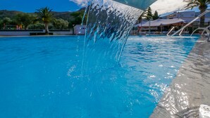 En swimmingpool med krystalklart, blåt vand, et vandfald og udsigt over det omkringliggende landskab med palmer og bygninger.