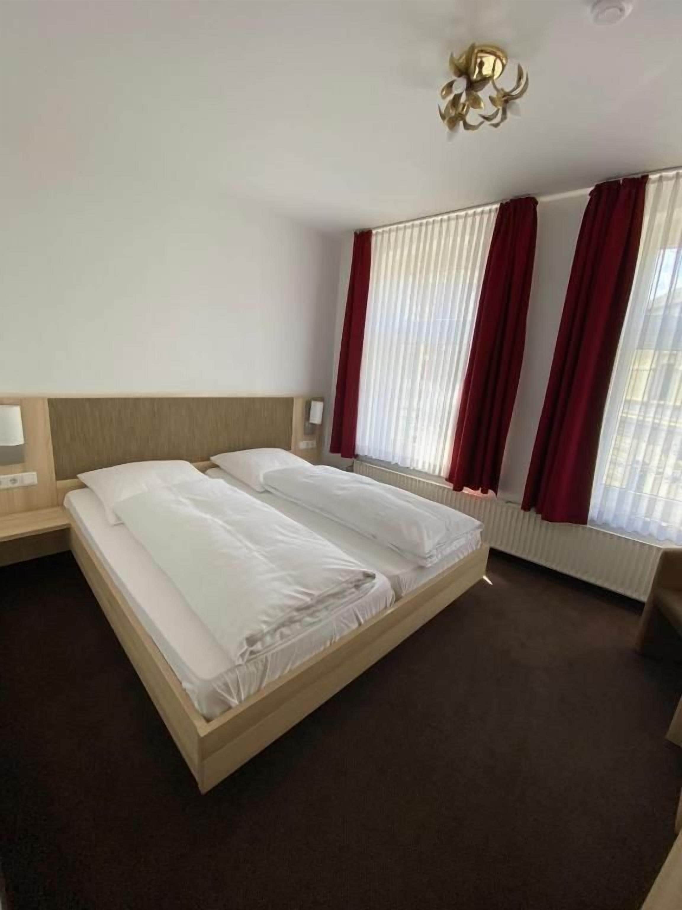 A hotel room with a double bed, a nightstand, a lamp, a window with curtains, and a radiator.