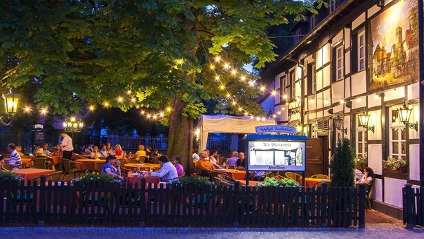 An outdoor dining area with tables and chairs, a signboard, and a building with a traditional facade. - Hotel Alt Westerholt (Herten)