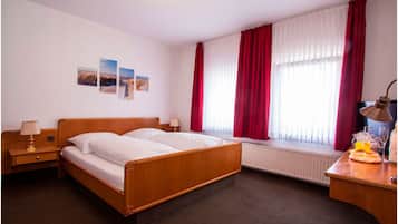 A hotel room with a wooden bed, two bedside tables with lamps, a radiator, and a window with red curtains.