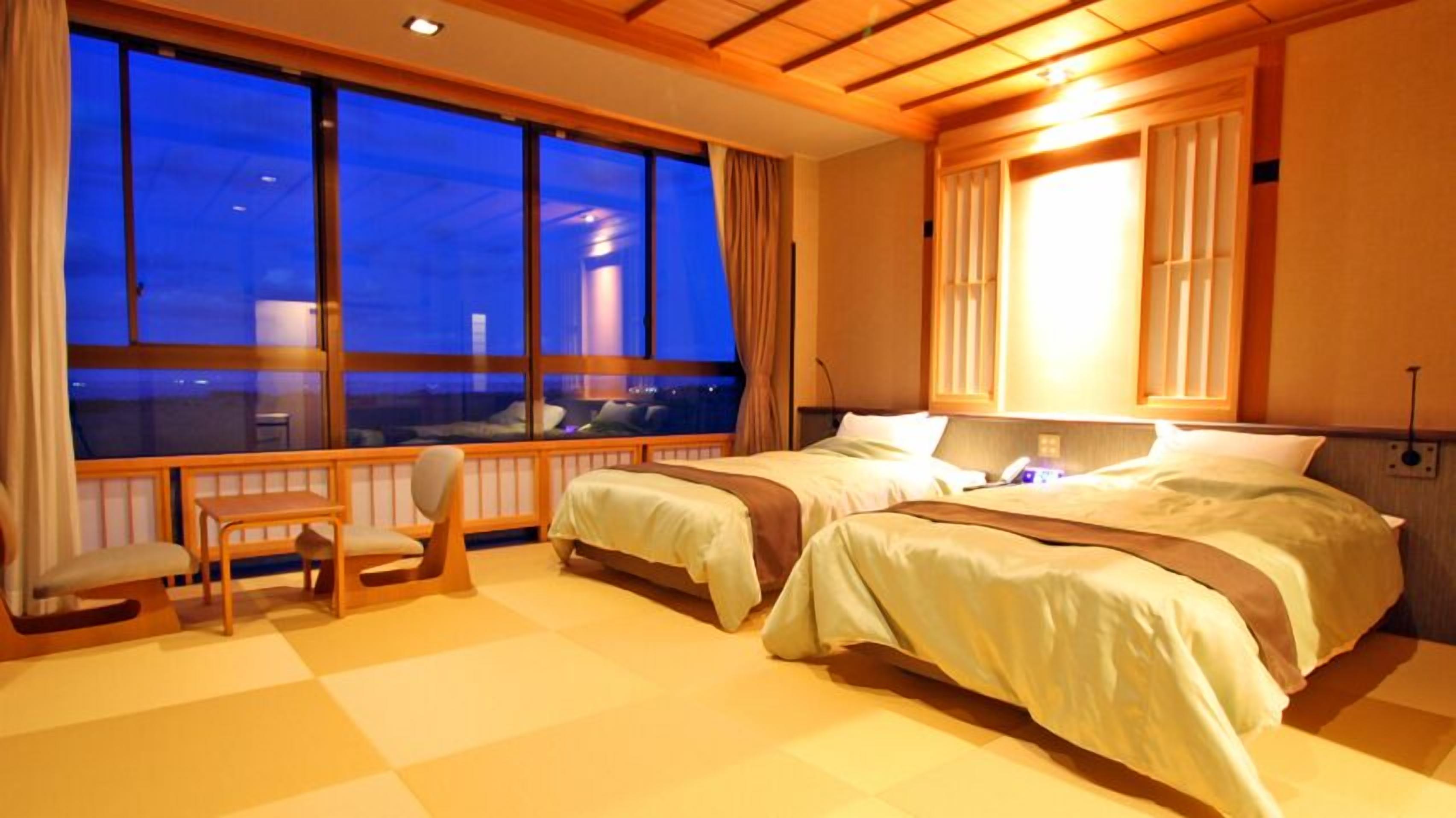 Habitación de hotel con dos camas, una silla, ventanas amplias y vista nocturna de la ciudad.