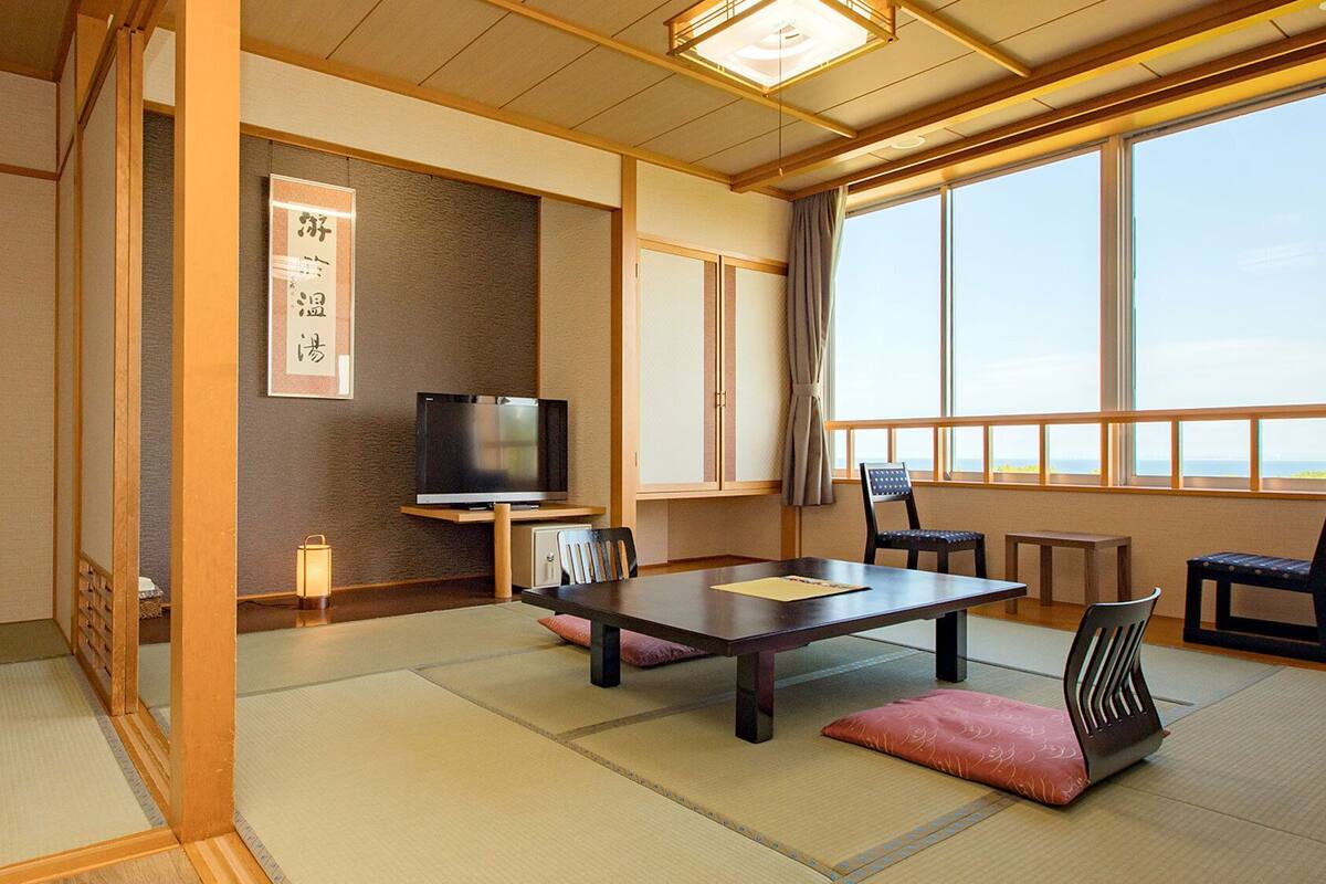Perinteinen japanilainen huone, jossa on tatamimatto, televisio, pöytä, tuolit ja suuri ikkuna, josta on kaunis näköala.