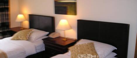 Een hotelkamer met twee bedden, elk met een kussen en een bedlampje. Daartussen staat een nachtkastje met een telefoon en een lamp.
