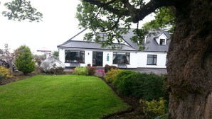 A white house with a grey roof, surrounded by a well-maintained lawn and various shrubs. - Mountain View Guesthouse (Oughterard)