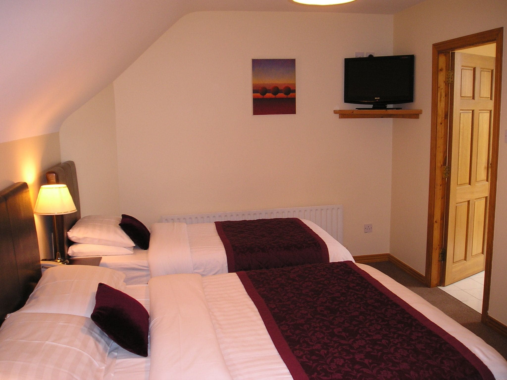 A hotel room with two beds, a television on a shelf, a lamp, and a door.