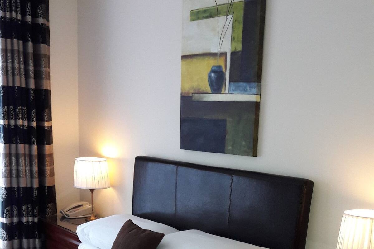 A neatly made bed with white linens and a dark headboard, flanked by two bedside tables with lamps. A painting hangs above the bed.