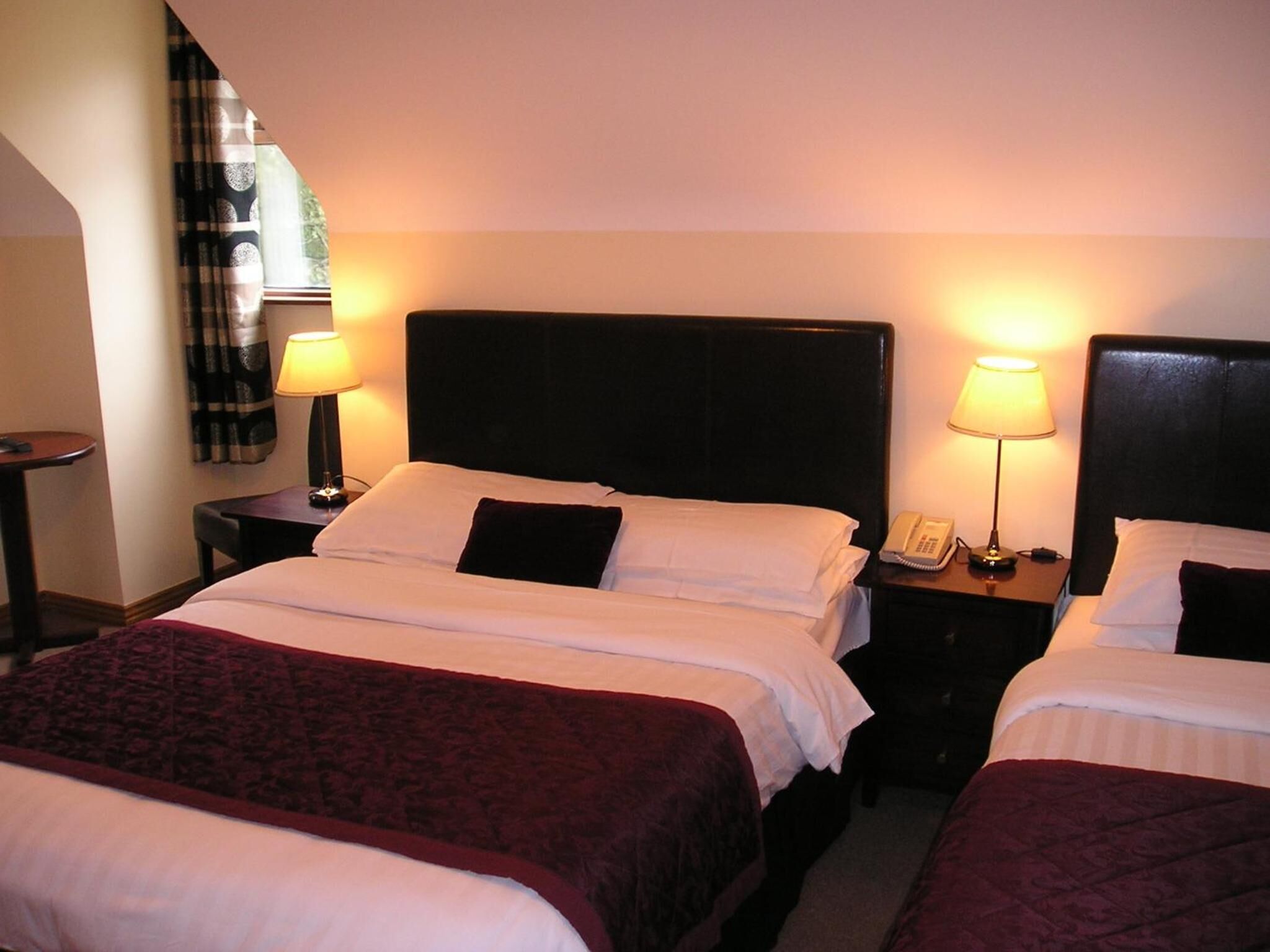 A hotel room with two beds, each with a black headboard, white linens, and a red throw blanket. There are bedside tables with lamps and a telephone.