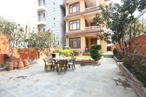 A courtyard with a stone floor, a round table with chairs, potted plants, and a building with balconies.
