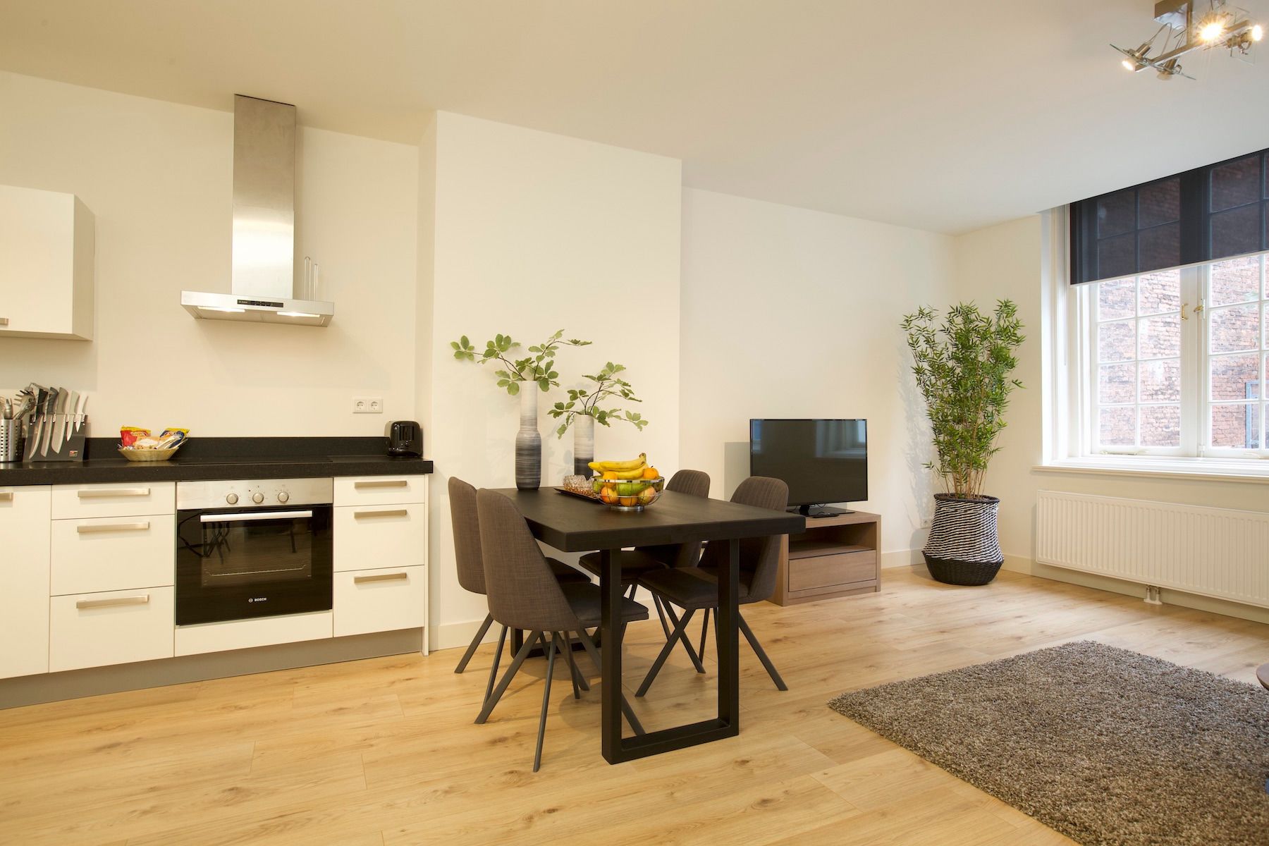 A modern kitchen with white cabinets, a black countertop, and a dining area with a table and chairs. There is a TV and a potted plant in the room.