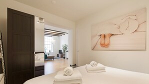 A hotel room with a large bed, two towels, a painting of feet, a dark door, and a view into the living area.