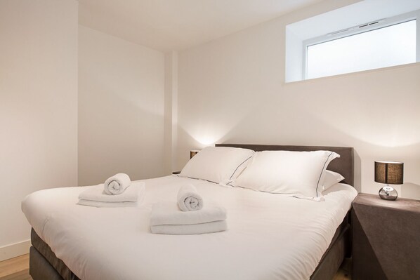 A neatly made bed with white linens and two rolled towels, a bedside table with a lamp, and a small window above the bed.