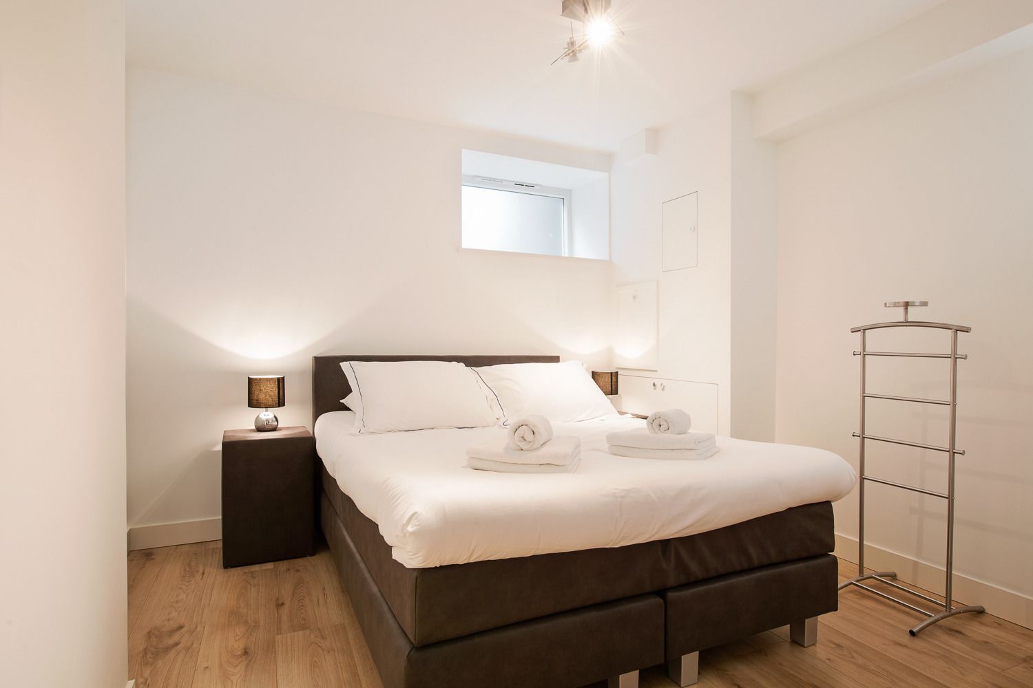 A modern bedroom with a large bed, bedside tables with lamps, a wardrobe, and a tall metal rack.