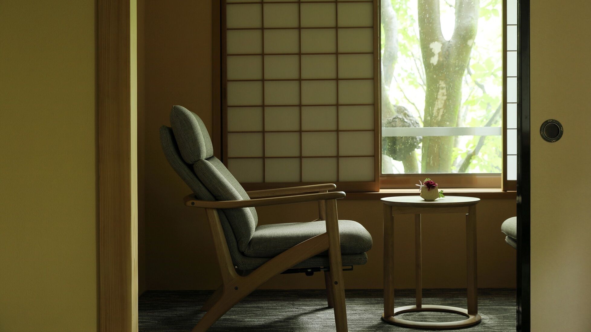 A room with a tatami mat wall, a wooden chair, a small round table with a vase, and a window with a view of trees.