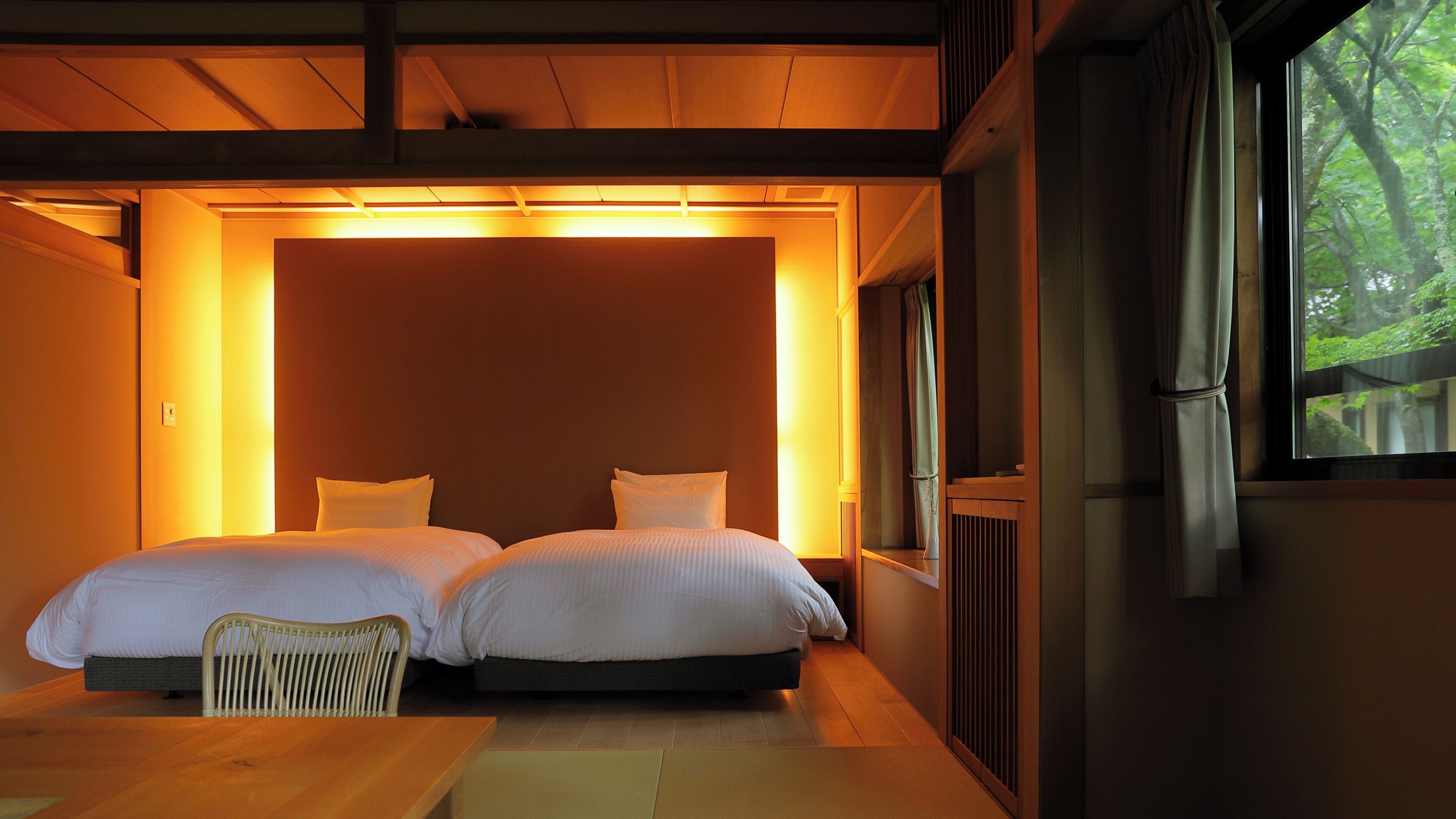A room with two beds, a wooden table, and a window with a view of trees.