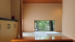 Una habitación minimalista con suelo de tatami, una puerta corredera que da a una vista de bosque y un estante de madera con un jarrón.