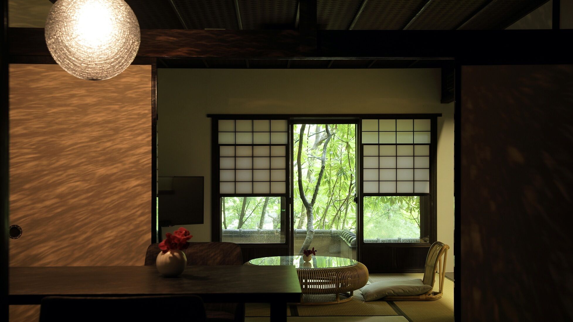 A traditional Japanese room with tatami flooring, a low table with a vase, a cushioned seating area, and a large window with a view of greenery.