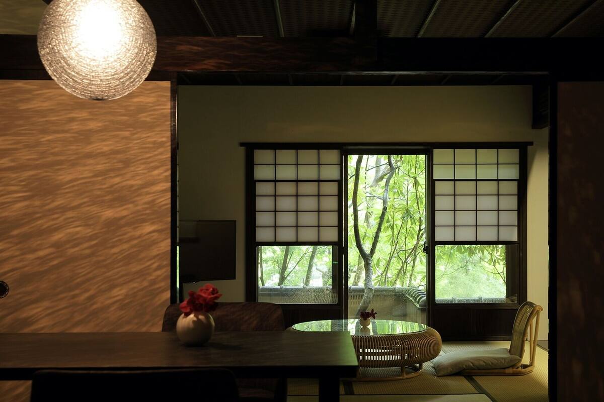 Een traditionele Japanse kamer met tatami vloeren, een lage tafel met een vaas, een zachte zithoek en een groot raam met uitzicht op groen.