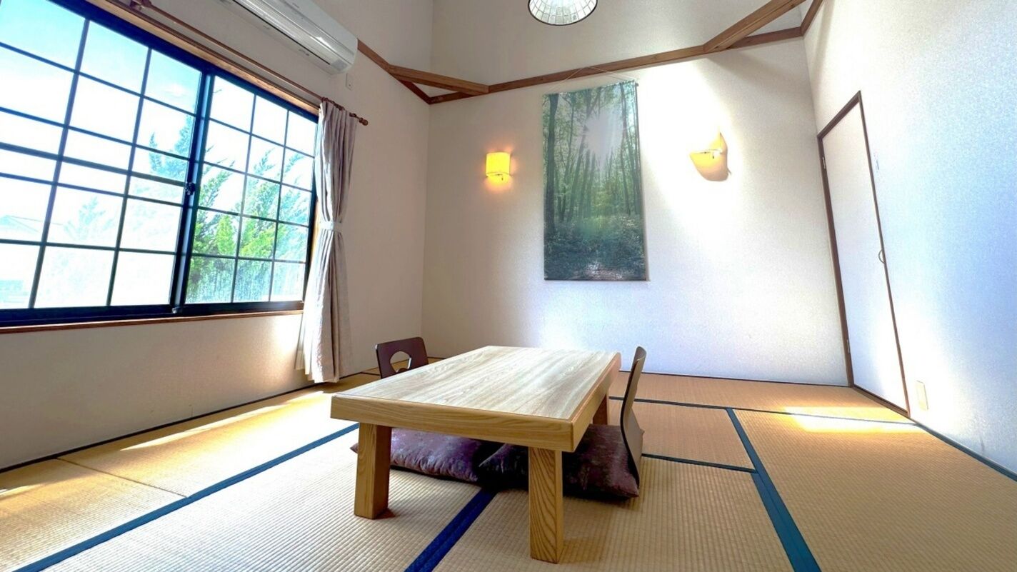 A tatami mat room with a low wooden table, tatami mats, a window with a view of trees, and two wall-mounted lights.