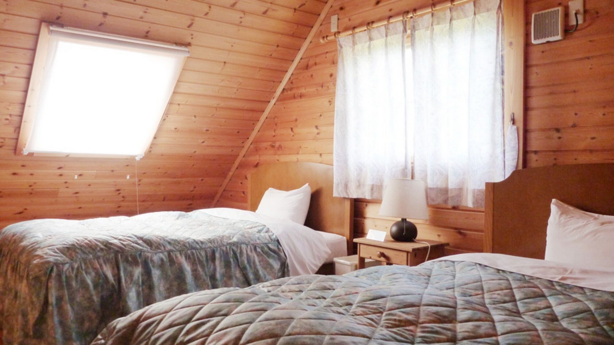 Une chambre de style cabane en bois, avec deux lits, une fenêtre avec des rideaux et une table de chevet avec une lampe.