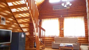 A log cabin interior with a wooden staircase, a television, a sofa, and a window with curtains.