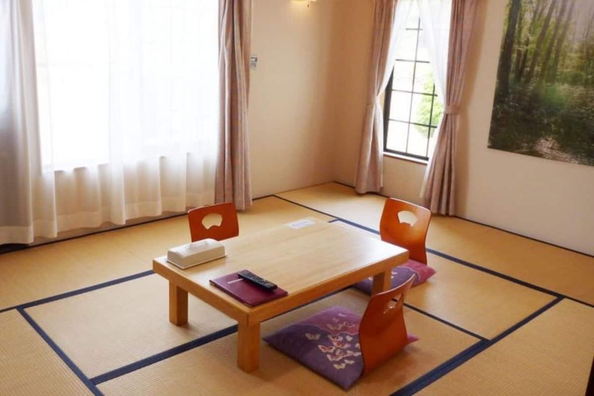 Une pièce avec un sol recouvert de tatamis, une table en bois, deux chaises, un lit et une fenêtre avec des rideaux.