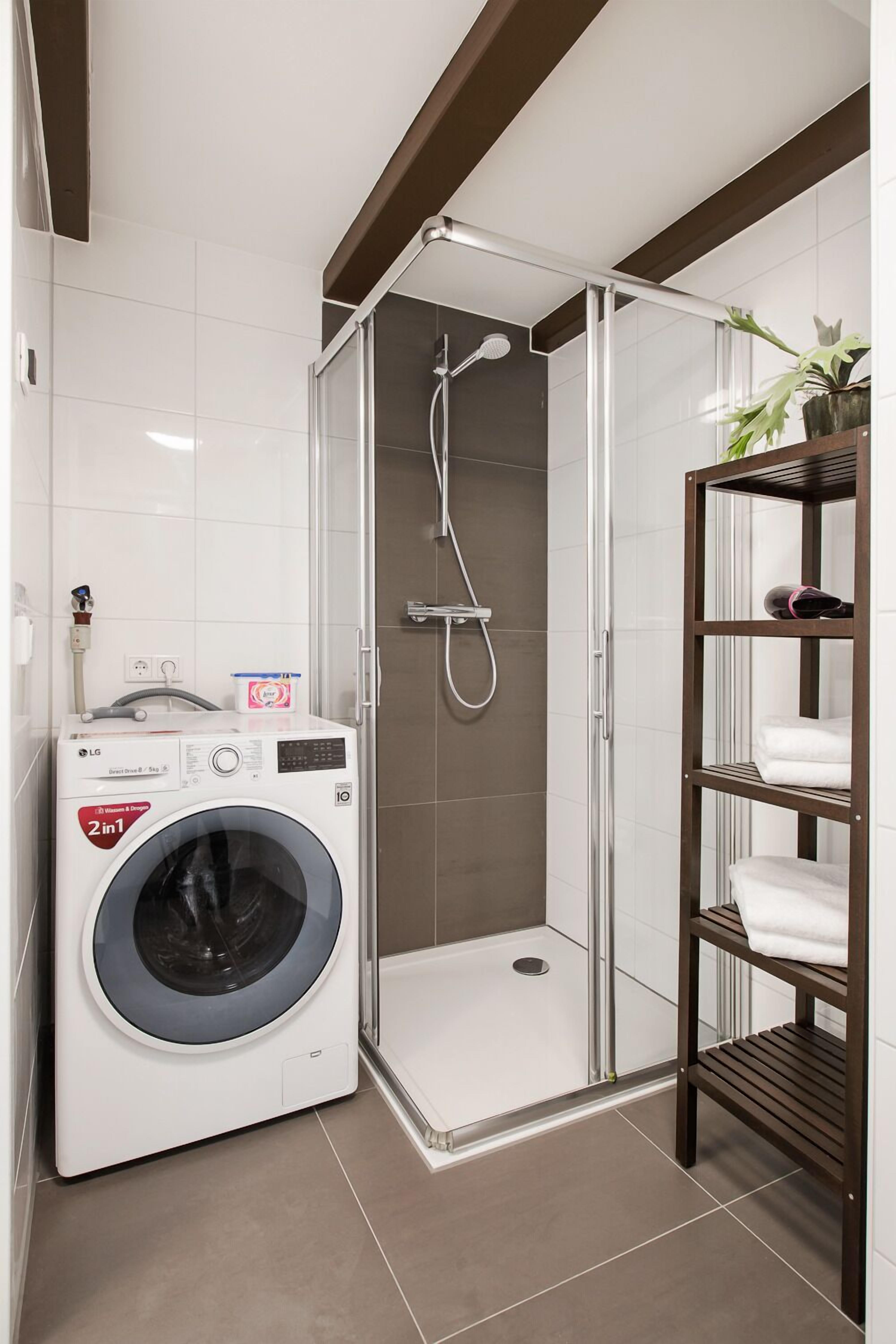 A compact bathroom with a washing machine, a shower enclosure, and a shelf with towels.