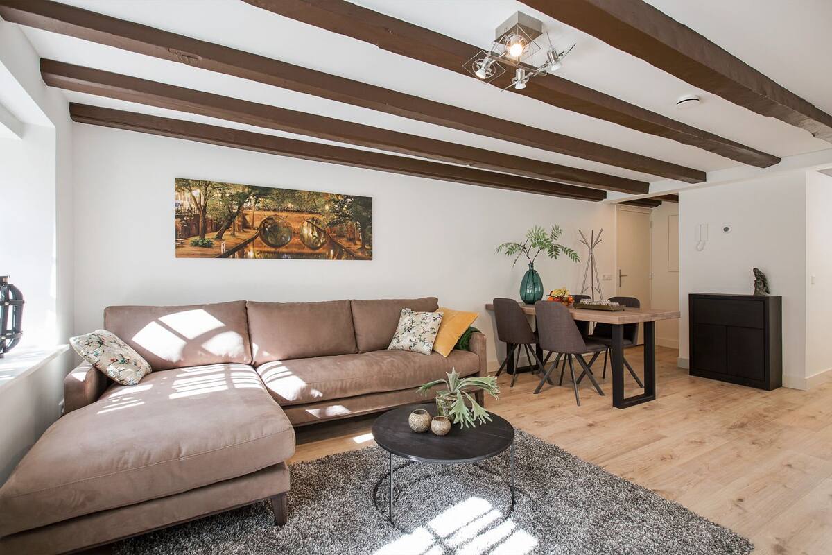 Uma sala de estar com um sofá marrom, uma mesa de jantar com cadeiras, uma mesa de centro preta e um quadro na parede.