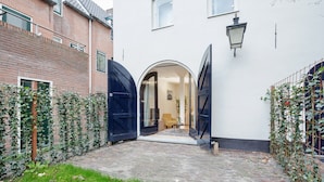 A courtyard with a brick building, two open black gates, and a white wall. - Canal Garden Apartment 11 (Utrecht)